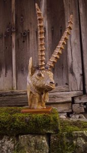 Hand-carved wooden ram statue with long spiraled horns beside a rustic wooden door, showcasing craftsmanship for wooden tables and doors.