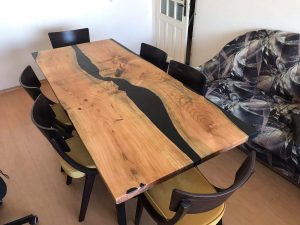 Handcrafted live-edge wooden dining table with a dark resin river, surrounded by chairs in a workshop, for a company that makes custom wooden tables and doors.