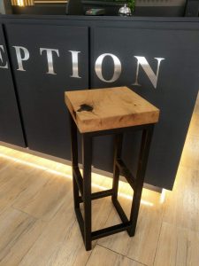 A square wooden-top stool with dark legs sits in front of a reception counter in a showroom, highlighting handcrafted wooden tables and doors.