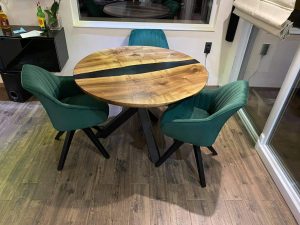 Round live-edge wooden dining table with a dark center inlay, surrounded by four green upholstered chairs in a showroom.