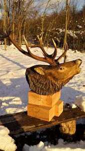 Handcrafted wooden deer head with large antlers resting on a wooden block on a bench in a snowy outdoor scene, reflecting the craftsmanship behind our wooden tables and doors.