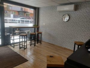 Bright woodworking showroom showcasing handcrafted wooden tables and stools by a large storefront window, with white brick walls and a dark display counter.