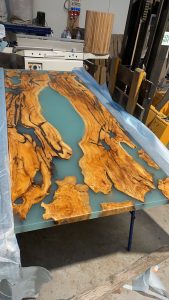 Handcrafted live-edge wooden table top with blue epoxy resin inlay, prepared in a workshop by a company specializing in wooden tables and doors.