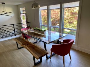 Bright dining area featuring a handcrafted wooden table with a live-edge bench and red chairs under large windows, highlighting the business’s wooden tables.