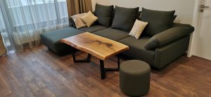 Modern living room with a handcrafted wooden coffee table, dark sofa with cushions, an ottoman, and hardwood floors, showcasing our custom wooden tables and doors.