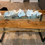 A woodworking studio with a live-edge wooden table top on a metal frame in the foreground and blue door-mold samples on a workbench in the background, highlighting custom wooden tables and doors.