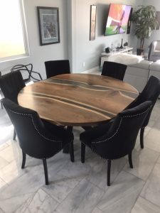 Round solid-wood dining table with natural grain and live-edge, surrounded by six black upholstered chairs in a modern dining room, showcasing handcrafted wooden tables and doors.