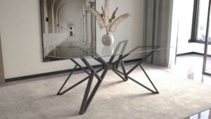 A glass-top coffee table with a black geometric X-shaped base sits in a living room, topped with a vase of dried pampas grass.