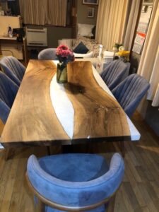 Live-edge wooden dining table with a white resin river, surrounded by blue upholstered chairs in a showroom.