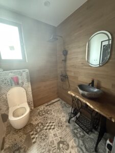 Bathroom interior with warm wood paneling and a handcrafted wooden vanity with a stone sink, a patterned tile floor, a toilet, a shower, and a round mirror.