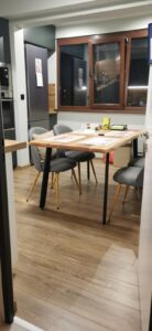 Showroom scene featuring a light wood dining table with gray upholstered chairs and dark wood framed doors and windows in the background, highlighting handcrafted wooden tables and doors.