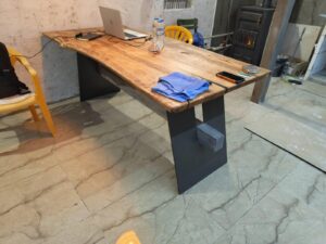 A wooden table with dark metal legs sits in a workshop, part of a furniture company that makes handcrafted wooden tables and doors.
